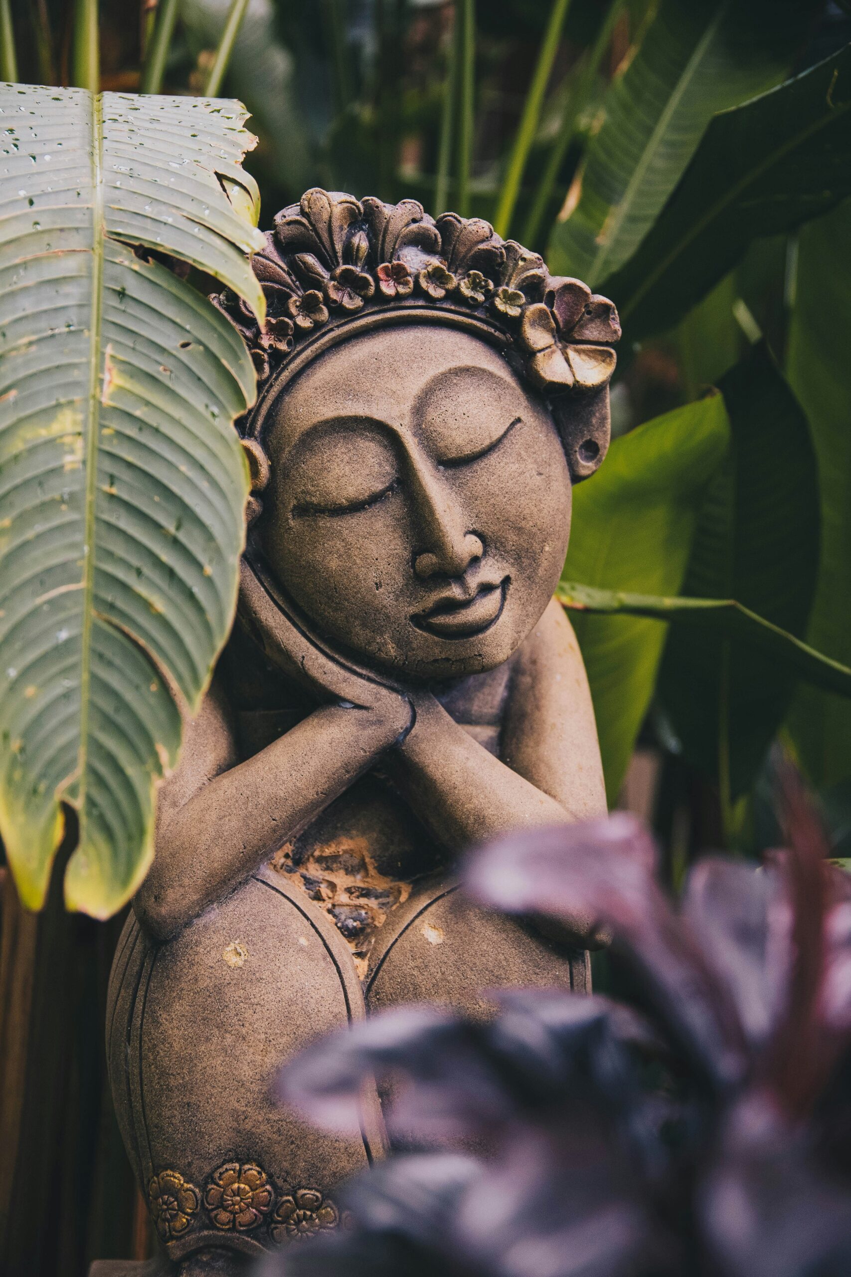 Serene-faced stone statue, eyes closed, looking deeply rested nested in low palm leaves.